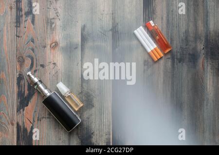 Schwarze E-Zigarette und süße E-Liquid gegen Tabakzigaretten mit Feuerzeug auf grauem Holzhintergrund überzogenem Rauch. Draufsicht. Stockfoto