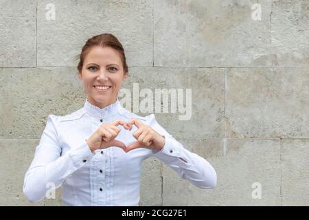 Attraktive lächelnde Frau in weißem Hemd zeigt Herz-Zeichen auf Marmor Backstein Wand Hintergrund mit Copy Space. Menschen Freundlichkeit Konzept. Stockfoto