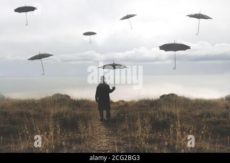 Einsamer Mann mit Schirm geht eine Straße hinunter in Richtung der see und surreale schwarze Schirme fliegen in den Himmel Stockfoto