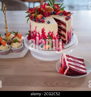 Große schöne rote Samtkuchen, mit Blumen und Beeren auf der Oberseite. Scheibe auf einem Teller, Dessert. Kuchen und Muffins, süße Urlaubssnacks. Stockfoto