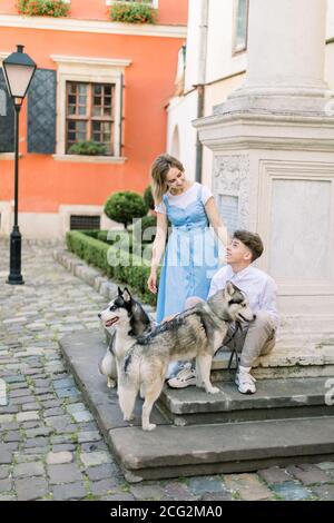 Glückliches Paar mit zwei Husky Hunde Spaß im Freien in der Stadt, sitzen auf der Treppe in der Nähe Denkmal im Hof mit orangen Gebäude auf der Stockfoto