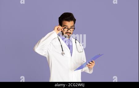 Junge indische Arzt in Uniform und Brille hält Zwischenablage an Violetter Hintergrund Stockfoto
