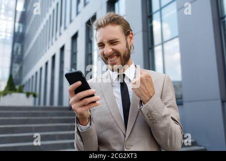 Aufgeregt und glücklich Geschäftsmann Gewinner hält ein Smartphone in Euphorie vom Sieg.Porträt eines lockigen jungen Geschäftsmann Manager in Ein Weiß Stockfoto
