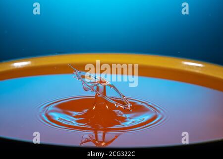 Zwei Tropfen orangefarbener Flüssigkeit fallen in den Becher, schlagen sich gegenseitig, dann wird die flüssige Oberfläche wie ein Pilz geformt Stockfoto