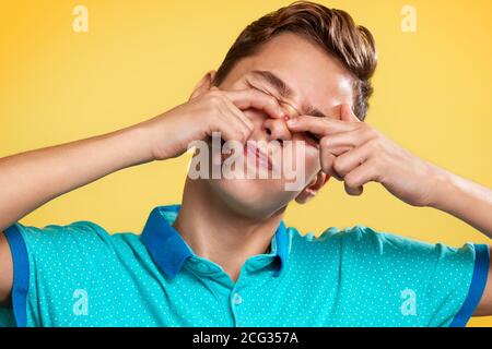 Kosmetologie, Dermatologie und Akne. Ein Teenager in einem blauen T-Shirt quetscht mit den Fingern einen Pickel auf seine Nase, die Augen sind vor Schmerzen geschlossen. Gelbe Rückenfarbe Stockfoto