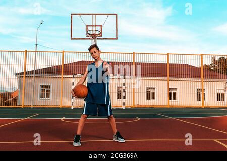 Sport und Basketball. Ein junger Teenager im blauen Trainingsanzug posiert selbstbewusst mit einem Ball, steht auf dem Spielplatz und winkt. Blauer Himmel und Bau Stockfoto