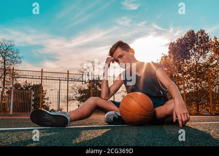 Sport und Basketball. Ein junger Teenager in einem blauen Trainingsanzug posiert mit einem Ball, während er auf einem Sportplatz sitzt. Blauer Himmel im Hintergrund. Speicherplatz kopieren. Stockfoto