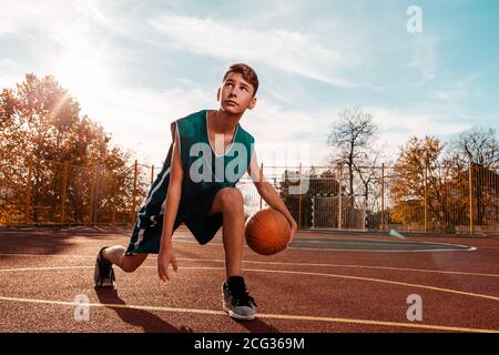 Sport und Basketball. Ein junger Teenager in einem grünen Trainingsanzug spielt Basketball und führt den Ball an. Blauer Himmel im Hintergrund und ein Sportplatz in t Stockfoto