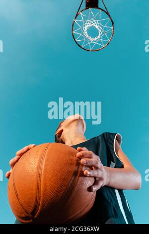 Sport und Basketball. Ein junger Teenager im schwarzen Trainingsanzug steht mit einem Ball in den Händen und bereitet sich darauf vor, den Ball in den Ring zu werfen. Blauer Himmel wi Stockfoto