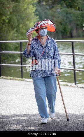 Eine stilvolle Frau in ihren Siebzigern geht für einen Übungsspaziergang mit einer chirurgischen Maske & passendem Hut & Geldbörse. In einem Park in Flushing, Queens, New York. Stockfoto