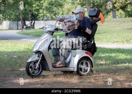 Ein älterer Mann, der eine Maske über dem Mund trägt, liest auf seinem dreirädrigen Motorroller an einem snady Fleck in einem Park eine vorausgesagt Sprachzeitung. In Queens, NYC. Stockfoto