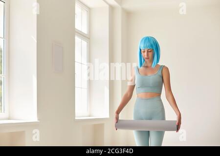 Junge Frau in eng anliegender Sportbekleidung steht im Fitnessstudio mit Yoga Matte in den Händen Stockfoto