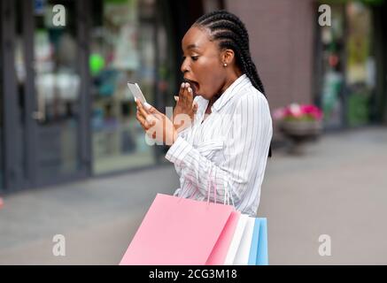 Ankündigung großer Rabatte in der mobilen Anwendung. Schockiert afroamerikanische Mädchen mit Taschen erhielt Nachricht auf dem Smartphone Stockfoto