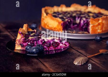 Rustikaler Shortbread-Kuchen mit Heidelbeeren in saurer Sahne Füllung auf einem Holzhintergrund mit Stück abgeschnitten auf Untertasse und Löffel. Stockfoto