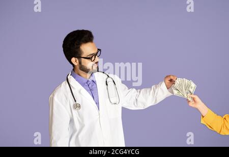 Korruption im Gesundheitswesen Konzept. Junge indische Arzt Geld von weiblichen Patienten über violetten Hintergrund Stockfoto