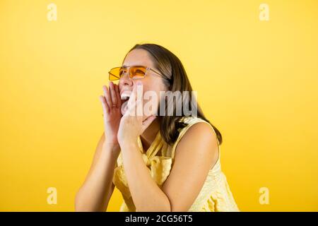 Junge schöne Frau trägt Sonnenbrille über isolierten gelben Hintergrund schreien Und schreiend laut zur Seite mit den Händen auf dem Mund Stockfoto