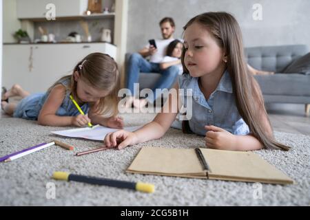 Kinder Schwestern spielen Zeichnung zusammen auf dem Boden, während junge Eltern zu Hause auf dem Sofa entspannen, kleine Mädchen Spaß haben Stockfoto