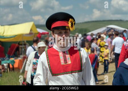 Ein junger Mann in russischer Volkstracht, der während des Volksfestes Karatag am Ufer eines großen Sees die Straße entlang läuft. Stockfoto