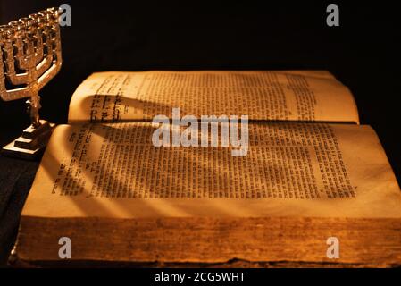 Hebräische Bibel und silberne Menora im Licht brennender Kerze auf dunklem Hintergrund. Schatten aus Menora auf offenen Seiten des jüdischen Gebetsbuchs in der Dunkelheit Stockfoto