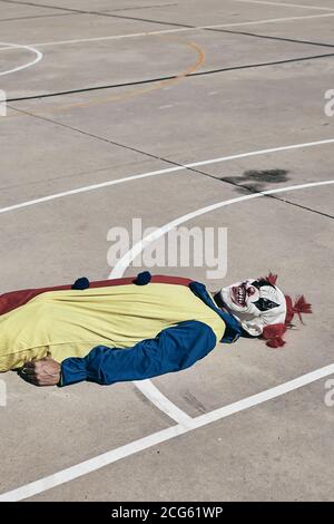 Ein gruseliger Clown, der ein gelbes, rotes und blaues Kostüm trägt und offen auf einem Basketballplatz im Freien liegt Stockfoto