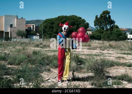 Ein gruseliger Clown, der ein gelbes, rotes und blaues Kostüm trägt, einen Haufen roter Luftballons in der Hand hält und auf einem freien Grundstück steht Stockfoto