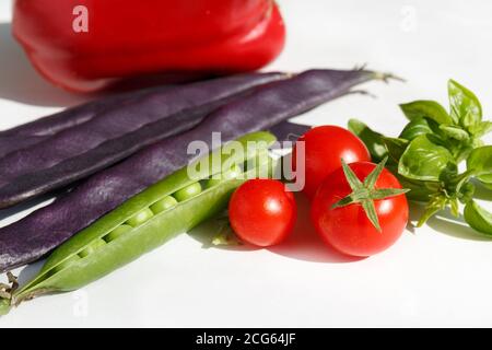 Kirschtomaten, grüne Basilikumblätter, eine offene Erbsenschote, violette Bohnenschoten und roter Paprika. Saisonales Gemüse auf weißem Hintergrund. Herbsternte Stockfoto