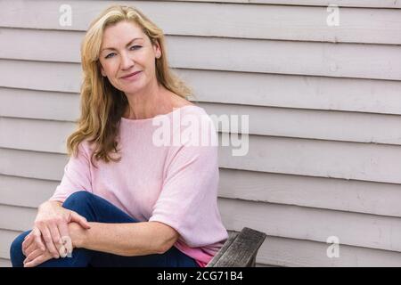 Outdoor-Porträt einer attraktiven mittleren Alters blonde Frau lächelnd Sitzen auf einem hölzernen Gartenstuhl Stockfoto