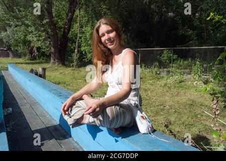 Ein rothaariges Mädchen sitzt mit gekreuzten Beinen auf einer alten blauen Holzbank vor einem Hintergrund von Bäumen. Stockfoto