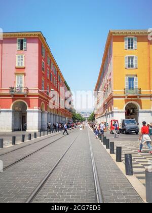 Menschen, die Fotos machen und in der Nähe von rot-ockerfarbenen Gebäuden mit Straßenbahn zwischen ihnen am Place Massena im Zentrum von Nizza, Frankreich, spazieren gehen. Stockfoto