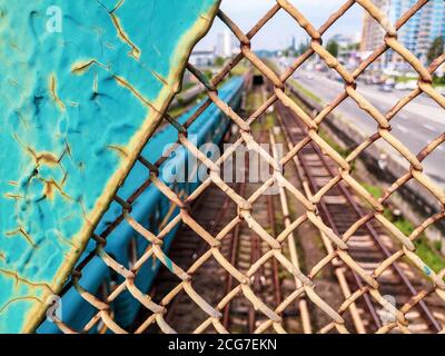 Blick durch den alten rostig lackierten rissigen Kettengliederzaun über die Eisenbahn mit blauer U-Bahn. Stadtbild von der Brücke. Stockfoto