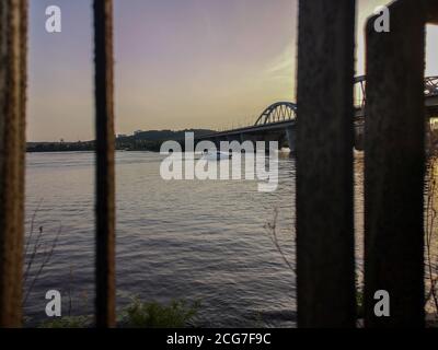 Blick durch das rostige Geländer über das Motorboot, das den Dnjepr unter der Darnytskyi-Brücke überquert. Blick vom linken Ufer der Stadt Kiew über das rechte Verbot Stockfoto