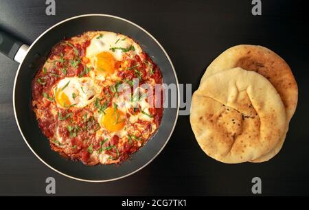 Shakshuka - pochierte Eier in Tomatensauce, Zwiebel, Pfeffer und Gewürze in eiserner Pfanne mit Pita Brot auf dunklem Holzgrund. Berühmte traditionelle arabische an Stockfoto