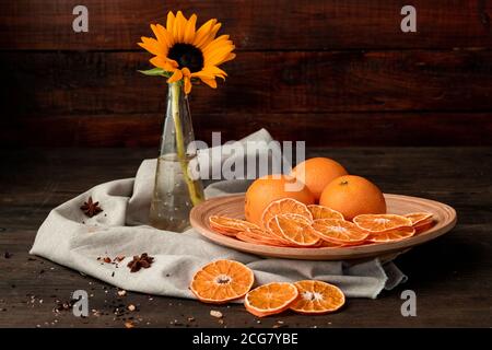 Schöne Zusammensetzung von frischen und getrockneten Orangen auf dem Teller und Sonnenblume in Vase auf Holzhintergrund Stockfoto