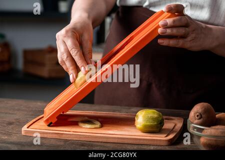 Nahaufnahme einer nicht erkennbaren Frau, die Kiwi auf einem Slicer über Holz schneidet Brett in der Küche Stockfoto