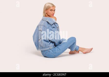 Freistil. Junge blonde Frau in Jeans und Jacke sitzen isoliert auf weiß zurückblickenden Kamera nachdenklich Stockfoto