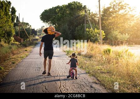 Kleines Mädchen, Fahrrad fahren, mit ihrem jungen Vater trägt einen großen halloween-Kürbis auf einer Landstraße bei Sonnenuntergang. Rückansicht. Stockfoto