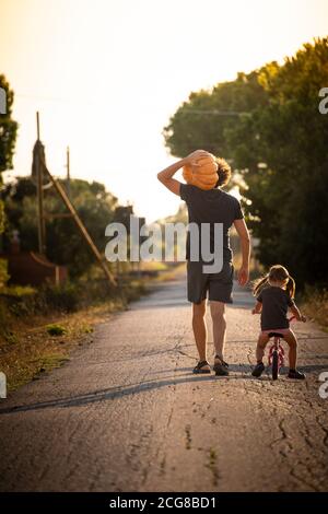 Kleines Mädchen, Fahrrad fahren, mit ihrem jungen Vater trägt einen großen halloween-Kürbis auf einer Landstraße bei Sonnenuntergang. Rückansicht. Vertikale Aufnahme. Stockfoto