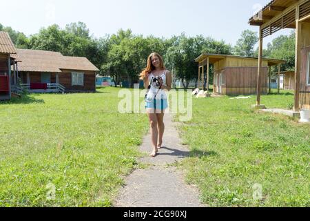 Ein fröhliches rothaariges Mädchen geht mit einer Kamera auf einem Weg auf einem Campingplatz, zwischen hölzernen Sommerhäusern, an einem sonnigen Tag. Stockfoto