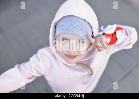 Kleines Mädchen mit blauen Augen in warmer Kleidung und Kapuze schaut auf und grüßt mit Geste. Stockfoto