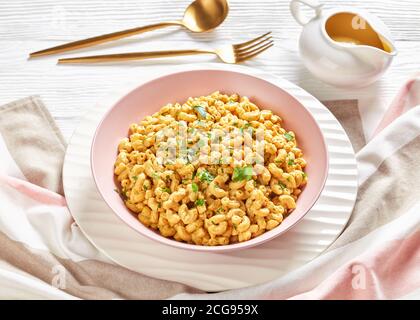 Cremiger veganer mac und Käse mit Hefesauce in einer rosa Schale auf einem weißen Holztisch mit goldenem Besteck, Landschaftsansicht von oben, close- Stockfoto