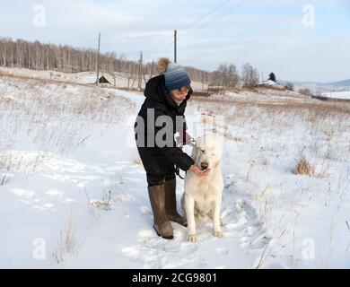 Eine Frau schleppt mit einer Delikatesse einen zentralasiatischen Schäferhund auf einer winterlichen Landstraße vor dem Hintergrund des Dorfes. Stockfoto