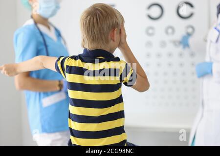 Boy sitzt mit dem Rücken beim Augenarzt Termin, schaut auf Tisch und beantwortet Fragen. Stockfoto