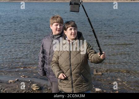 Mutter und Sohn werden auf einem Handy mit einem speziellen Stock auf dem Hintergrund des Flusses Jenissei fotografiert. Stockfoto