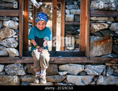 Junge Sherpa Mädchen mit Bandana Stirnband sitzt auf dem Fenster Rahmen von unter Bau Haus und lächelt an der Kamera In der abgelegenen Manaslu Region von Nepa Stockfoto