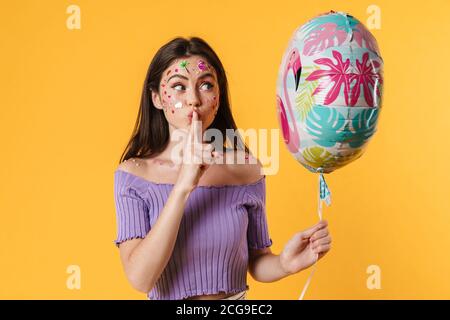 Bild der jungen Frau mit Aufklebern auf Gesicht hält Ballon Und zeigt Stille Geste isoliert auf gelbem Hintergrund Stockfoto