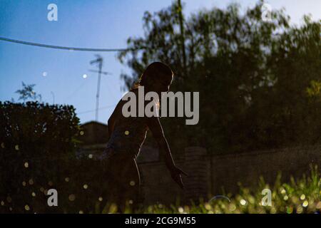 Silhouetten von Kindern, die mit dem Wasser spielen, das kommt Aus den Sprinklern einen Garten mit dem zu bewässern Sonne gegen das Licht, das im dr reflektiert wird Stockfoto