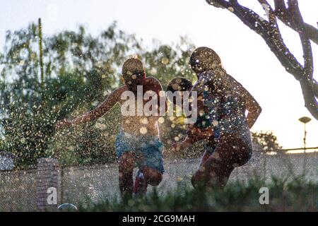 Silhouetten von Kindern, die mit dem Wasser spielen, das kommt Aus den Sprinklern einen Garten mit dem zu bewässern Sonne gegen das Licht, das im dr reflektiert wird Stockfoto