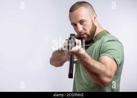 Ernst kaukasischen bärtigen Mann in lässigen grünen T-Shirt mit Ziel gekleidet Bei der Kamera mit Scharfschützengewehr, das isoliert auf Weiß zielt Stockfoto