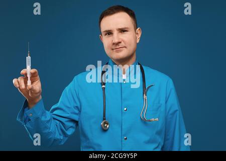 Junger männlicher Arzt in einer blauen medizinischen Uniform, der in der Kamera schaut und eine medizinische Spritze zur Injektion hält, isoliert auf blauem Hintergrund. Saisonal Stockfoto