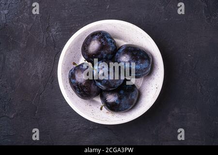 Frische reife Pflaumenfrüchte mit Wassertropfen in Holzschüssel auf dunklem Steingrund, Draufsicht Stockfoto